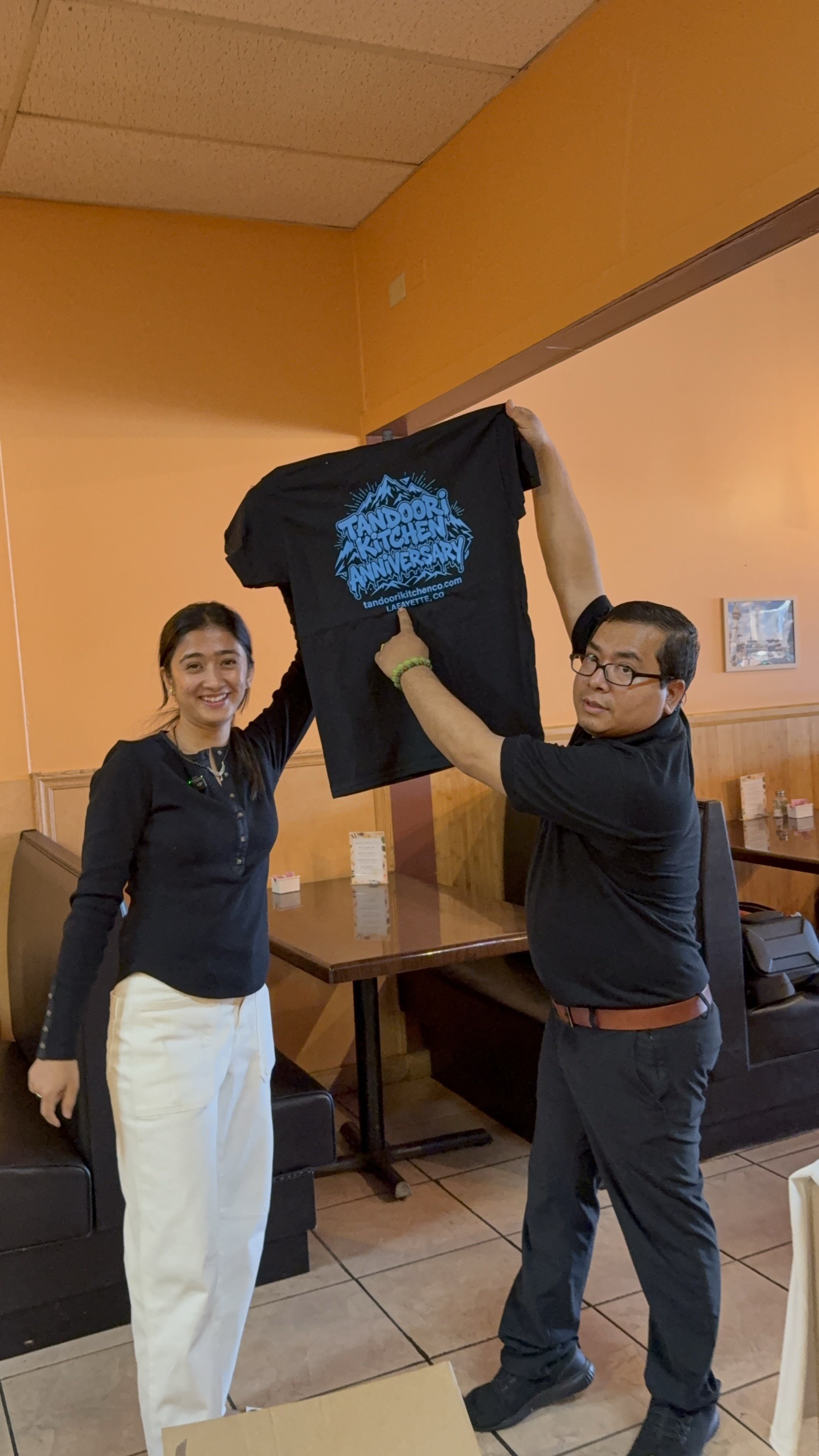 Two Tandoori Kitchen team members hold up the black anniversary tee.