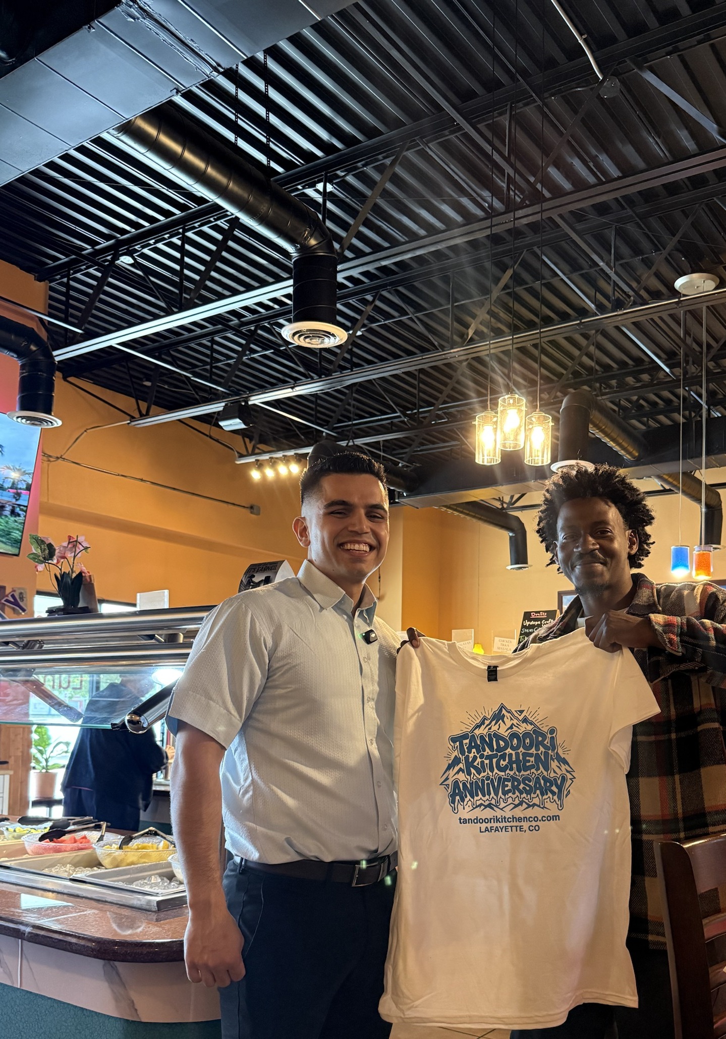 A guest and a Tandoori Kitchen server hold up the first anniversary tee in the dining room.