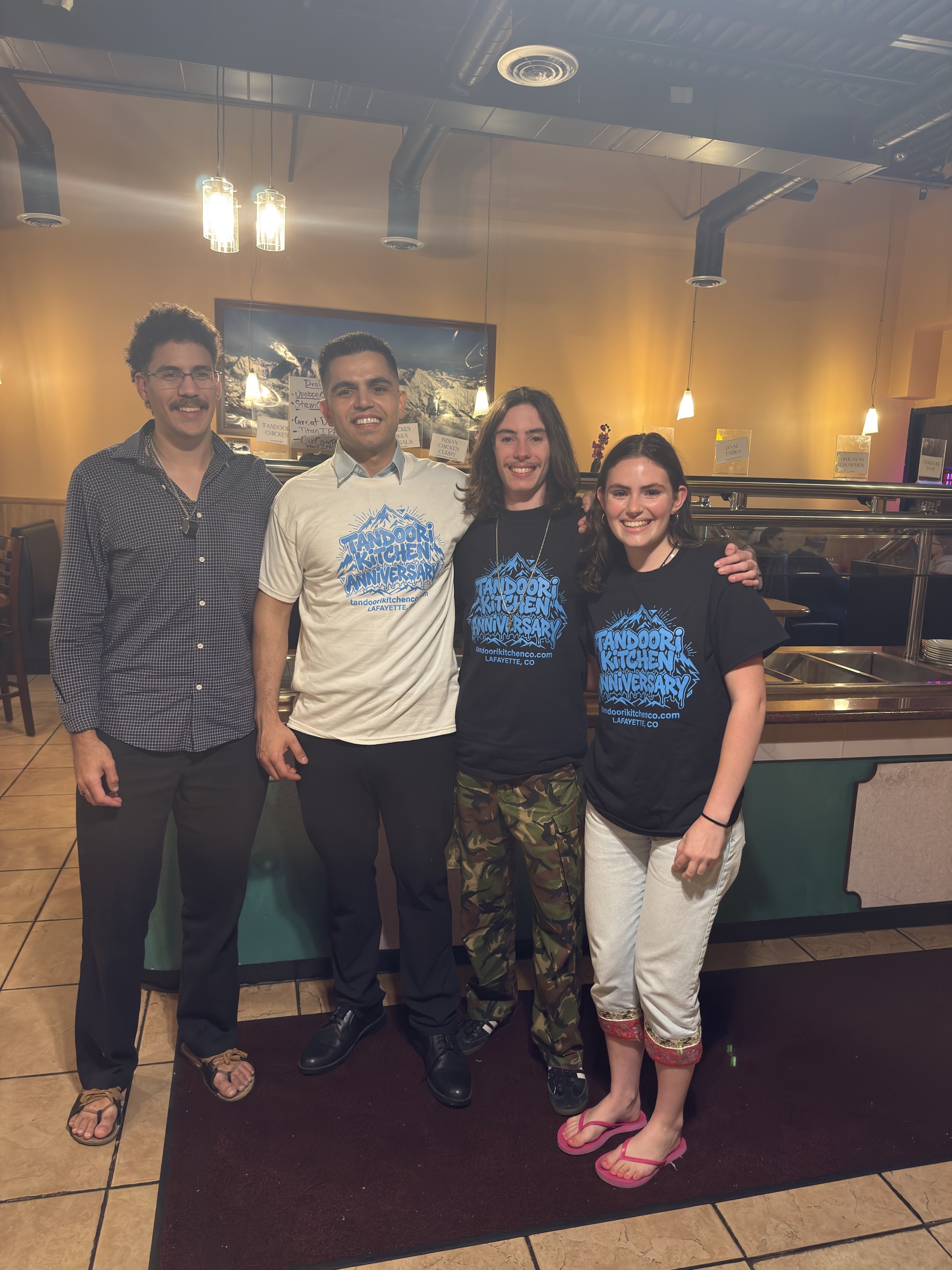 Guests wearing Tandoori Kitchen anniversary tees pose with a team member in the dining room.