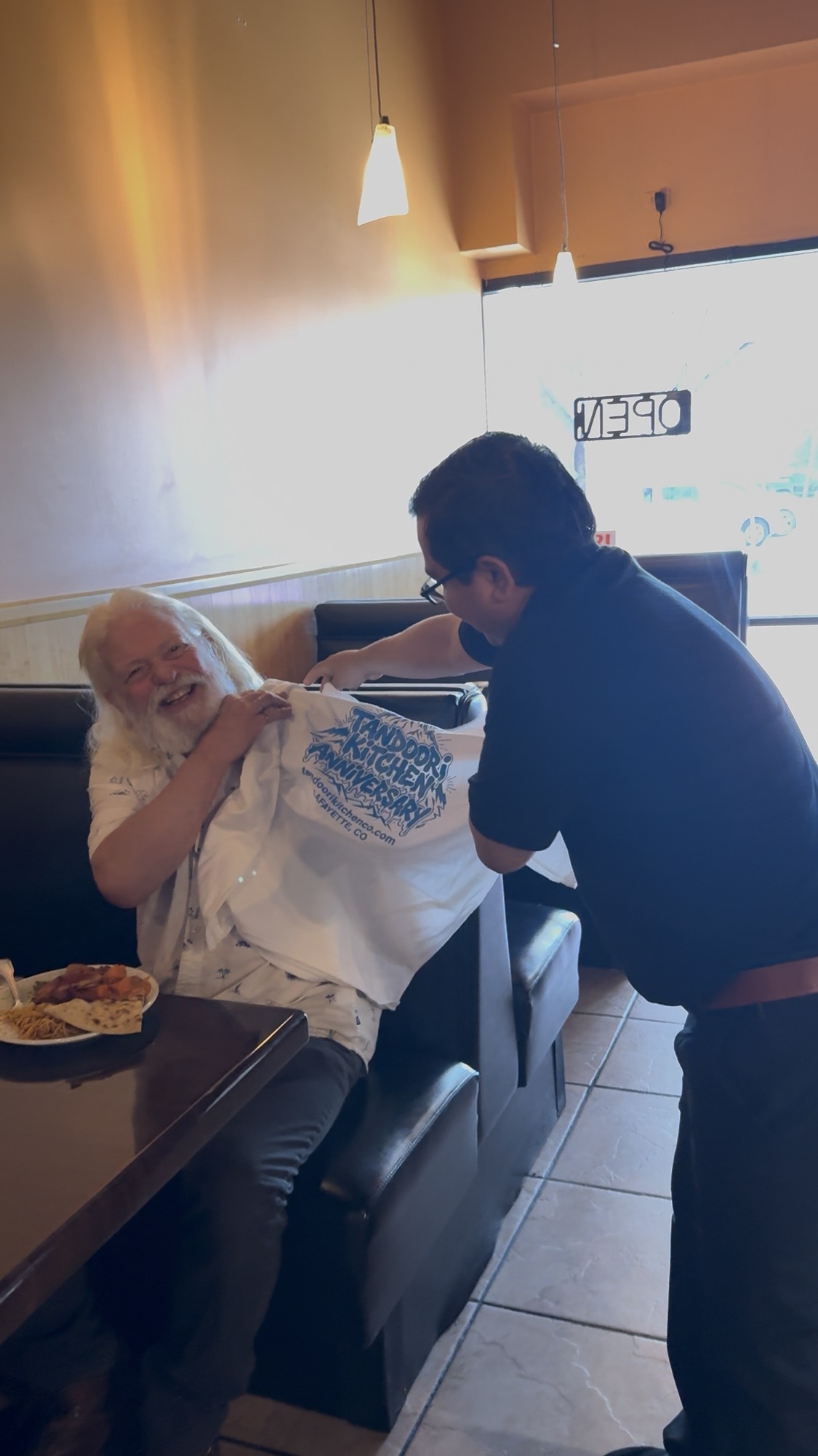 A longtime guest laughs as a Tandoori Kitchen team member hands over an anniversary tee at his table.