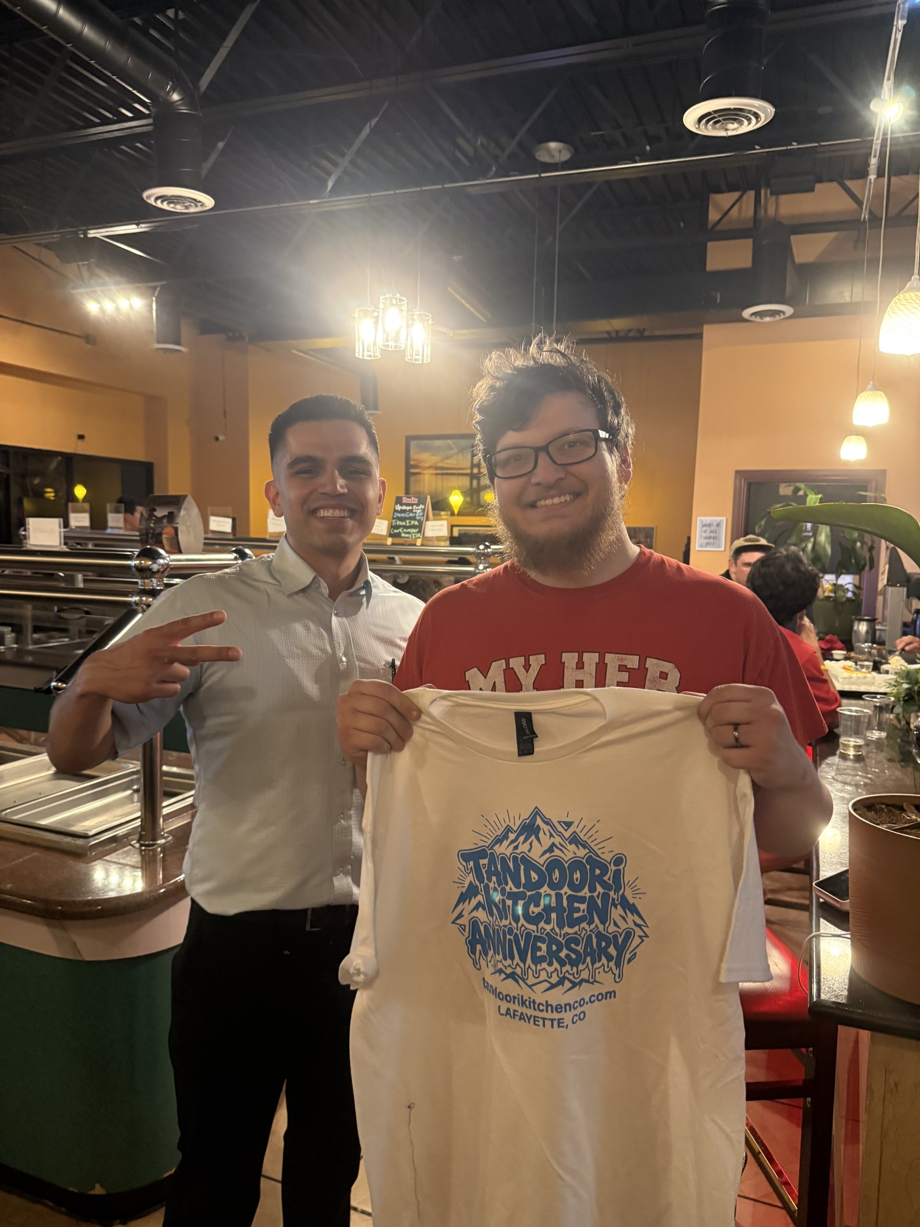A guest holds up a white anniversary tee next to a Tandoori Kitchen team member.